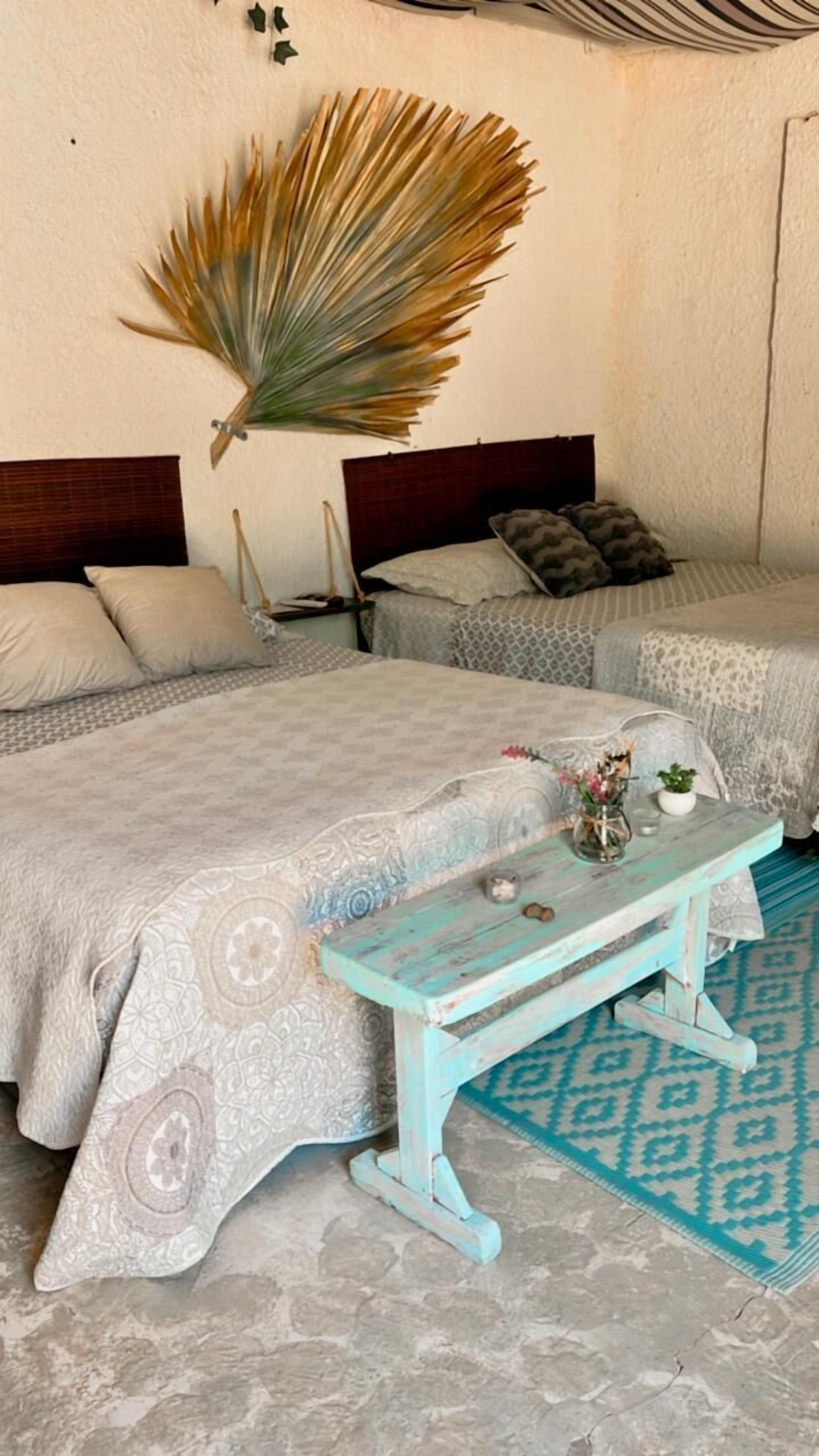 Une chambre avec deux lits, une petite table sur laquelle est posé un vase, et un grand éventail décoratif accroché au mur.