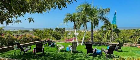 An outdoor seating area with chairs and tables, surrounded by greenery and a clear view of the ocean.