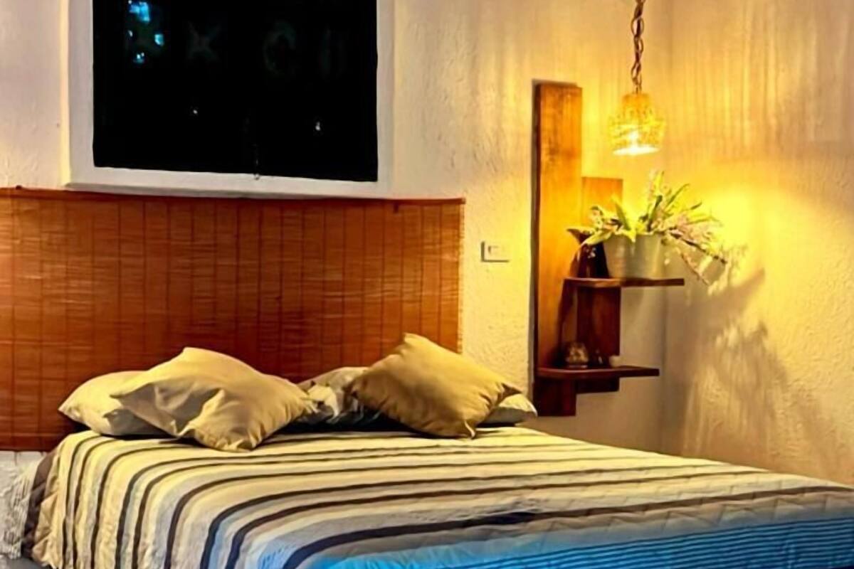 Ein Schlafzimmer mit einem Bett, einem Holz-Kopfteil, einem Fenster mit Jalousien, einer hängenden Pflanze und einem farbenfrohen Teppich.
