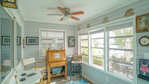 Una habitación con ventilador de techo, sillas de madera, una mesa y vistas al exterior a través de amplios ventanales.