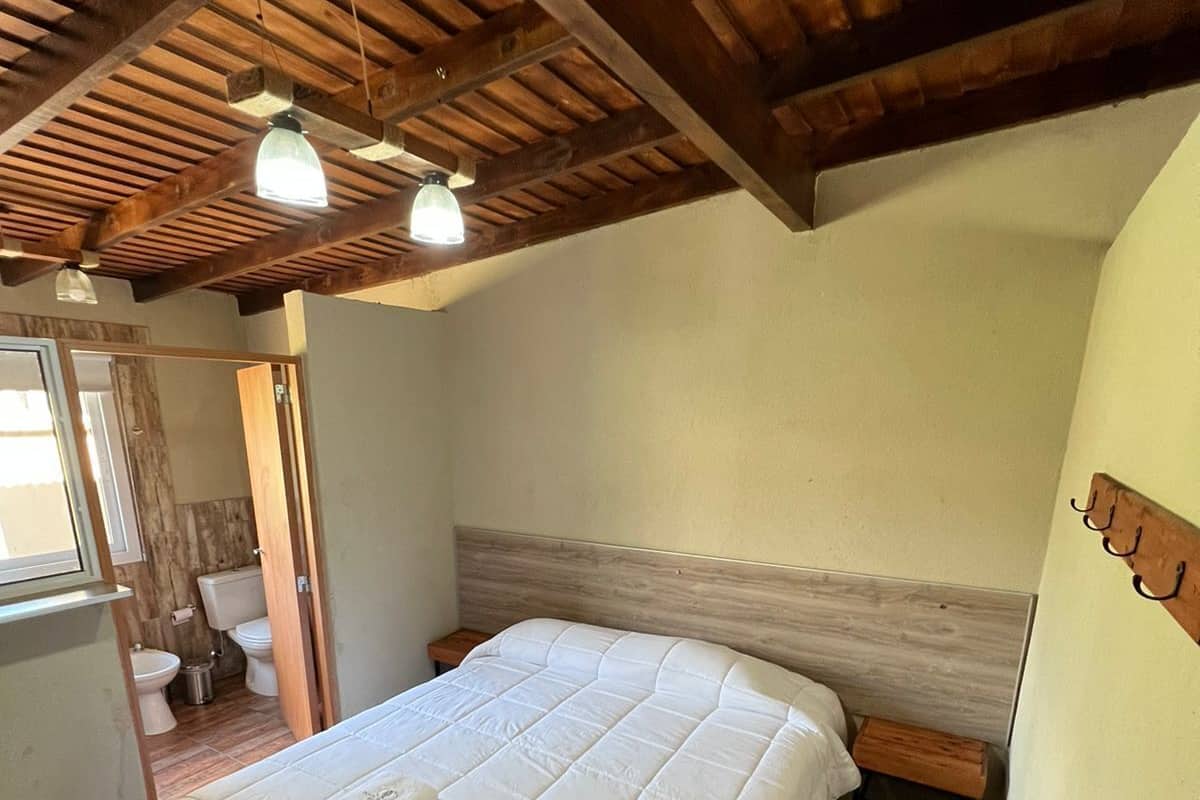 Un dormitorio con techo de madera, una cama con edredón blanco, una mesita de noche y una ventana con cortinas.
