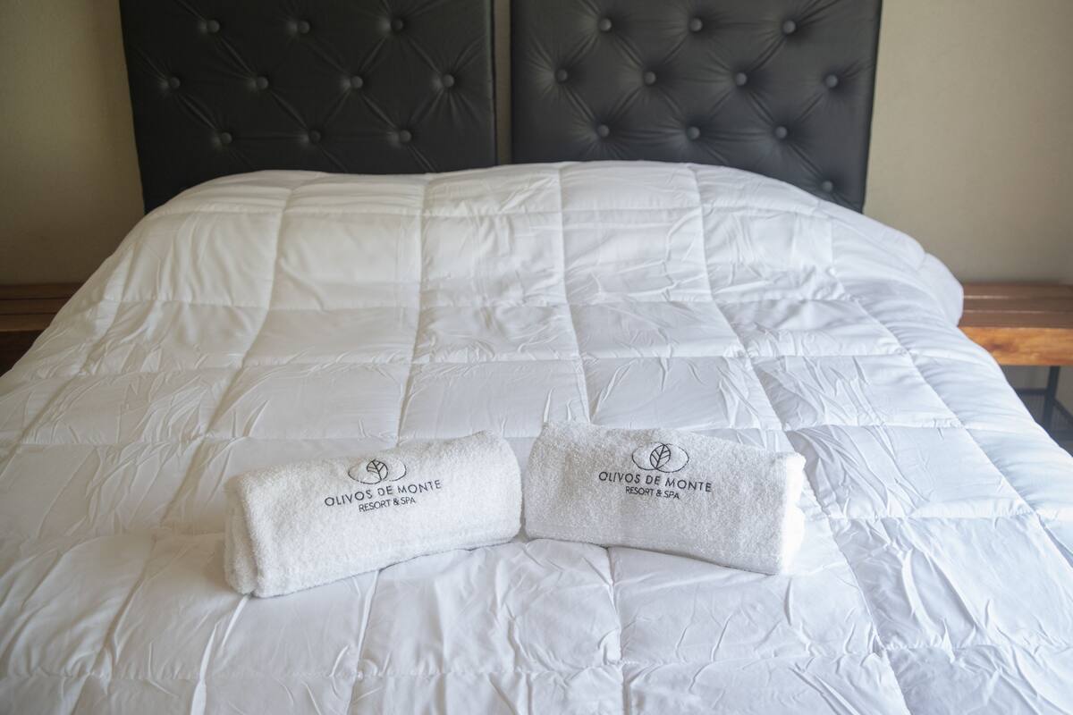 Una cama con una colcha blanca acolchada y dos toallas con el logotipo de “Olivos de Monte Resort & Spa” sobre una mesita de noche de madera.