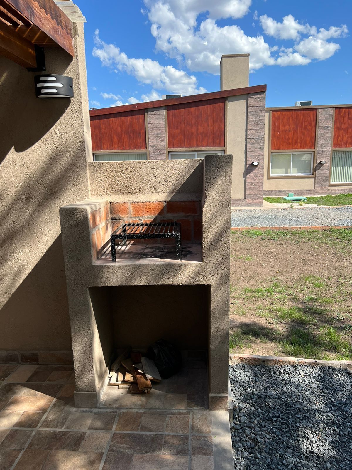 An outdoor barbecue area with a stone grill, a fire pit below, and a view of a modern building in the background.