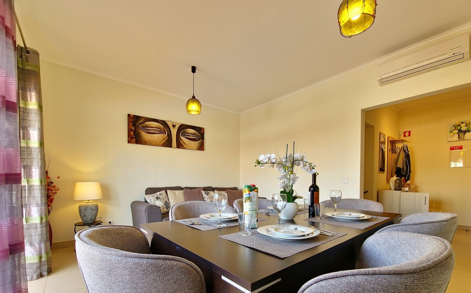 A dining area with a rectangular table set for four, surrounded by grey upholstered chairs. There is a wall-mounted artwork above the table and a hanging pendant light.