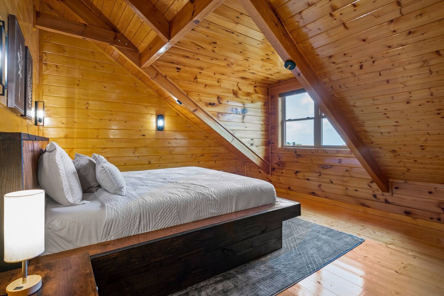 Une chambre mansardée chaleureuse avec un plafond en pente en bois, un lit avec des oreillers, une table de chevet et une fenêtre.