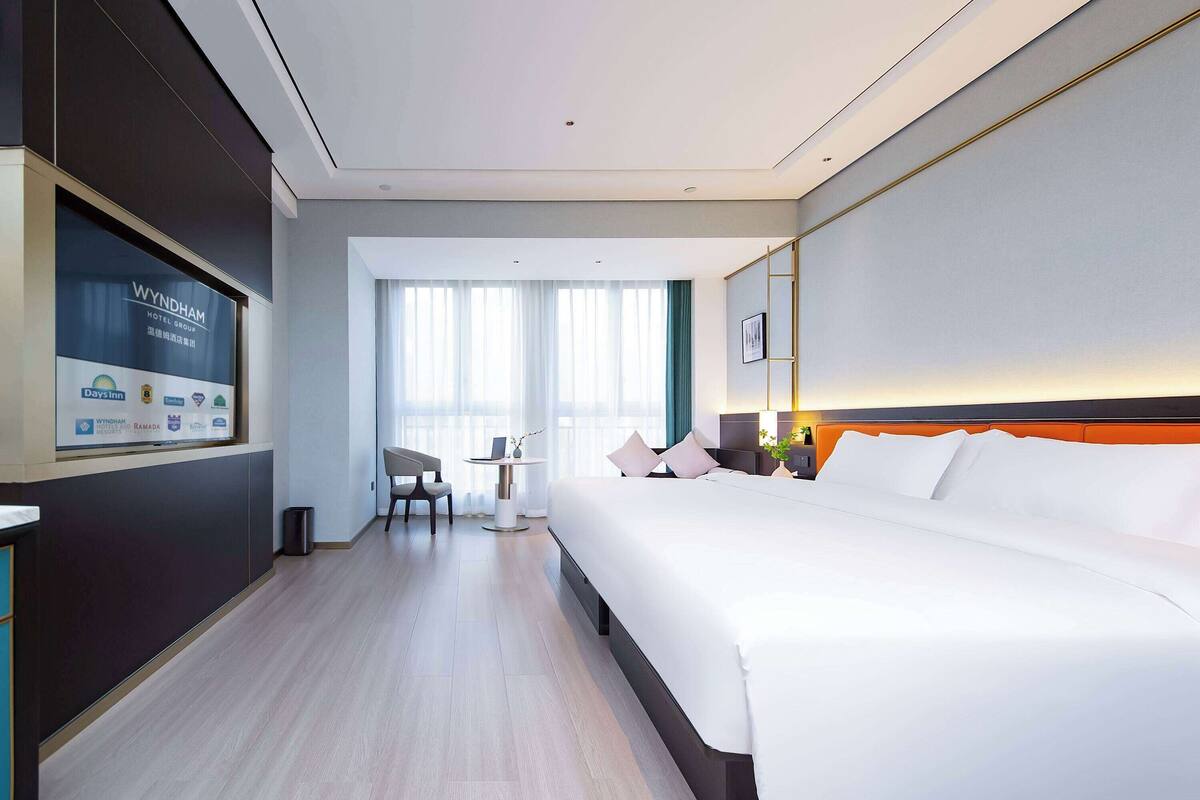 A hotel room with a large bed, a desk, a chair, and a TV displaying hotel information.