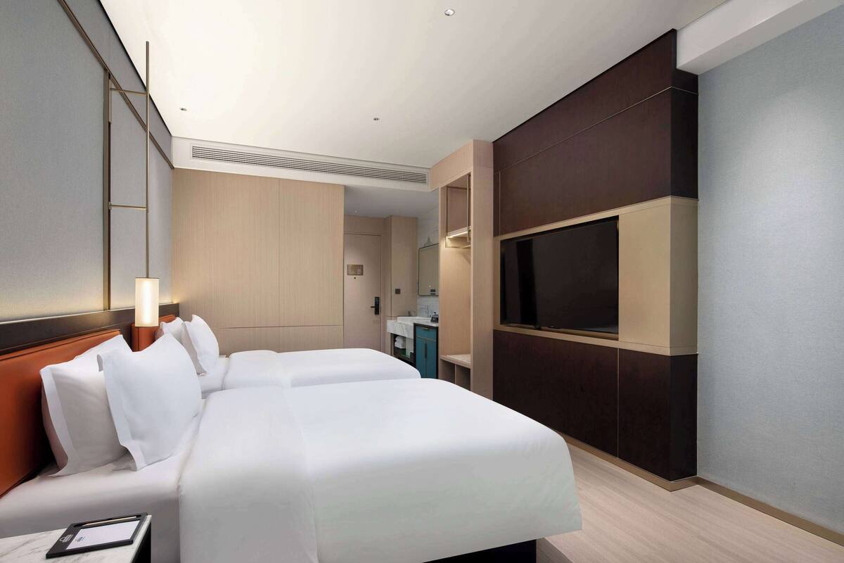 A hotel room with two beds, a TV, a desk with a lamp, and a bathroom visible in the background.