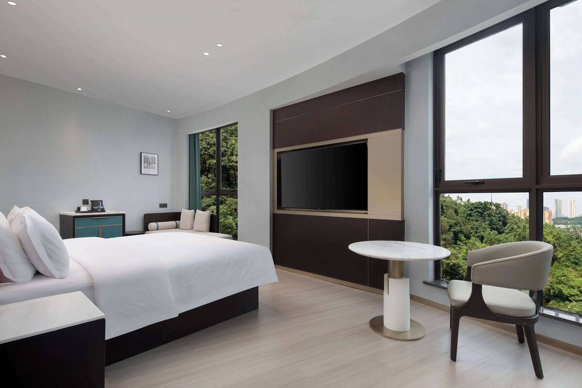 A modern hotel room with a large bed, a TV mounted on the wall, a small table, and a chair.