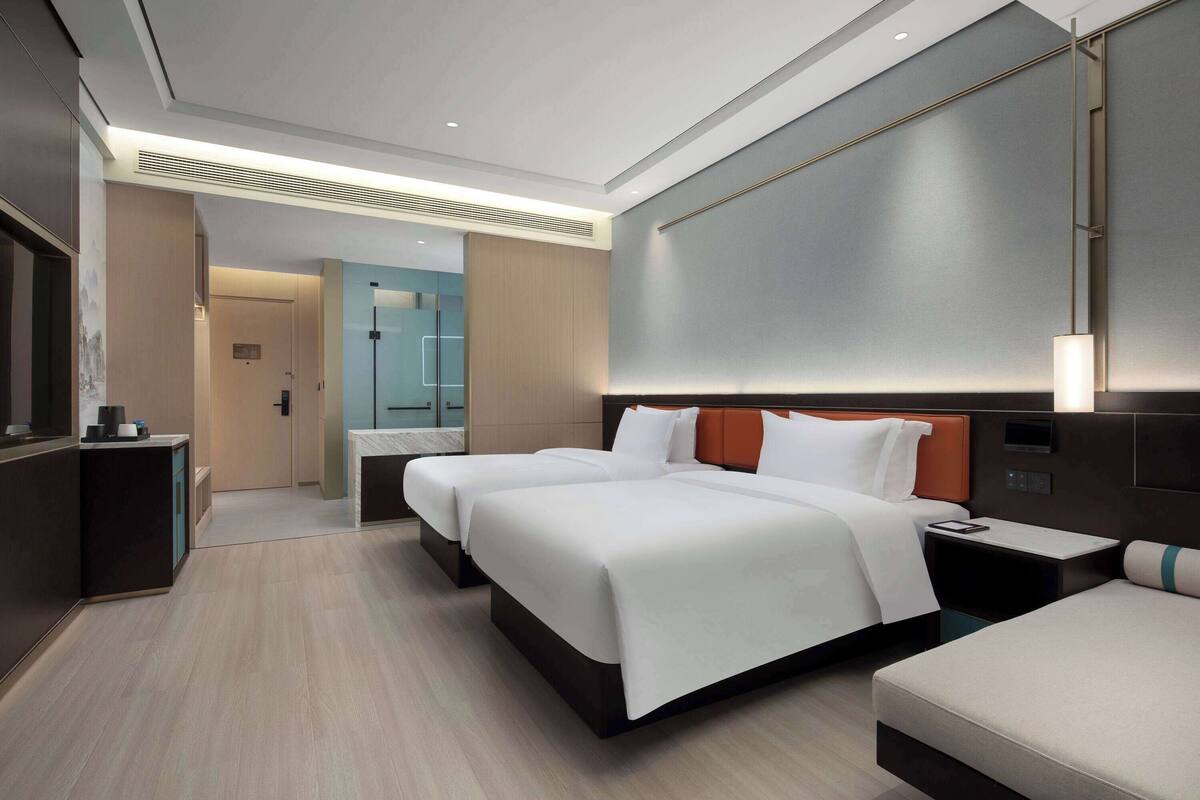 A modern hotel room with two beds, a desk, and a bathroom visible in the background.