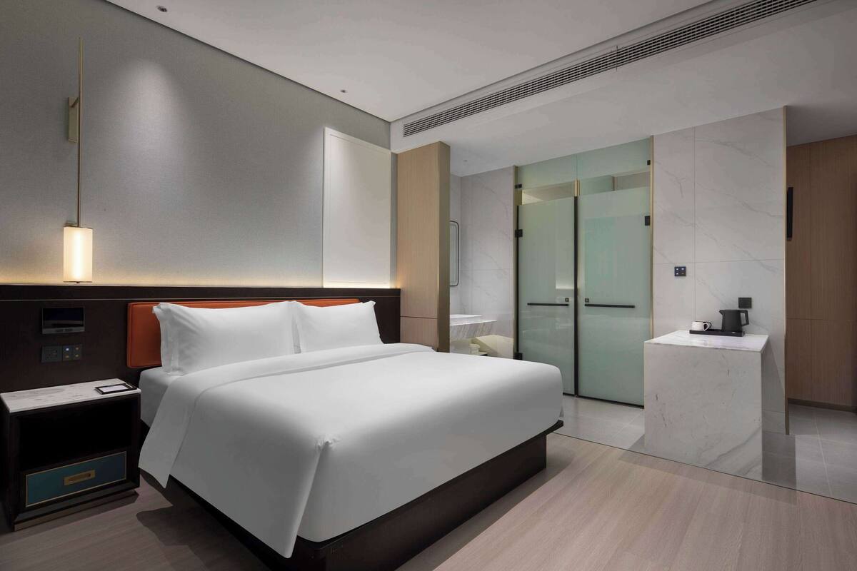 A modern hotel room with a large bed, a bedside table, a nightstand, a mirror, a small desk with a chair, and a glass door leading to a bathroom.