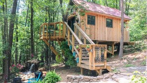 Uma cabana de madeira com escadas e um pequeno alpendre, rodeada por árvores e uma cachoeira.