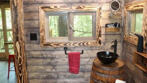 A rustic bathroom with wooden walls, a large window, a black basin on a barrel, and a red towel hanging.