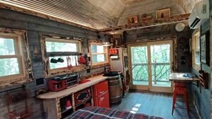 A rustic cabin interior with a kitchen area, a dining table, and large windows.