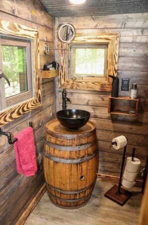 A rustic bathroom with wooden walls, a wooden barrel sink, a mirror, and a window. - Greers Ferry Lake Treehouses (Edgemont)