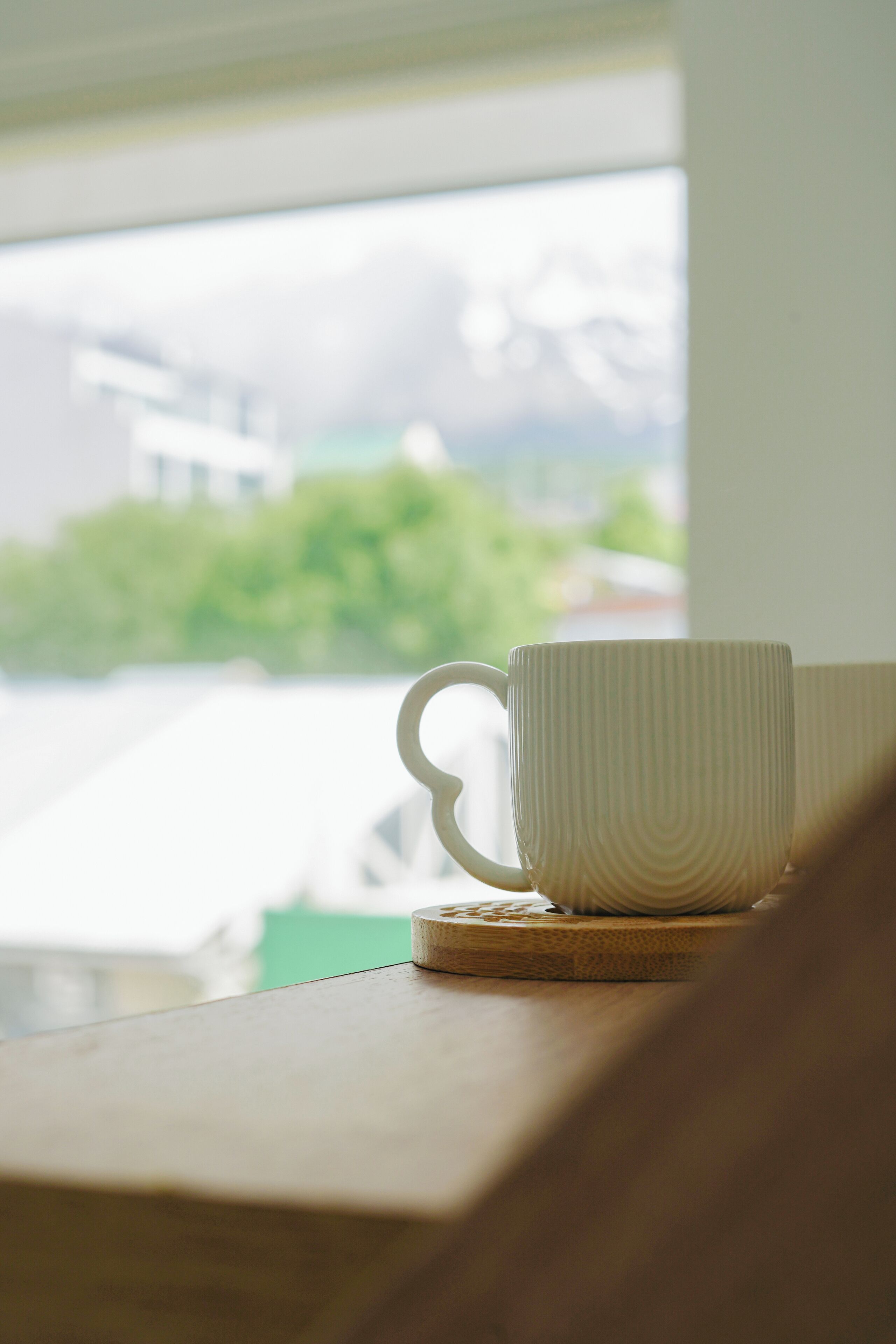 A white cup with a handle on a wooden surface, with a blurred view of greenery and buildings outside a window.