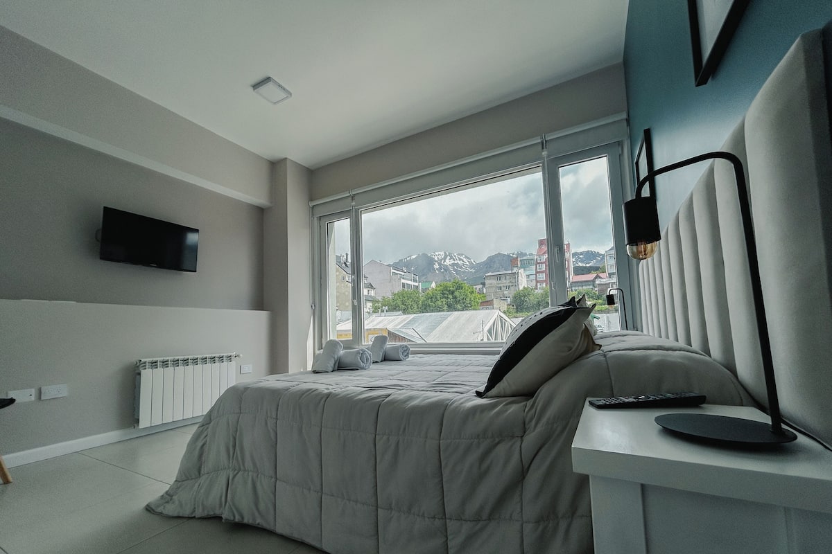 Quarto moderno com uma cama grande, uma mesinha de cabeceira, uma TV na parede e uma janela com vista para edifícios e montanhas.