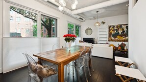 Un comedor moderno con una mesa de madera y sillas transparentes, ventanas amplias y un cuadro en la pared.