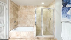 Un baño con bañera, ducha envuelta en vidrio, paredes de azulejos color beige y un cuadro abstracto enmarcado.