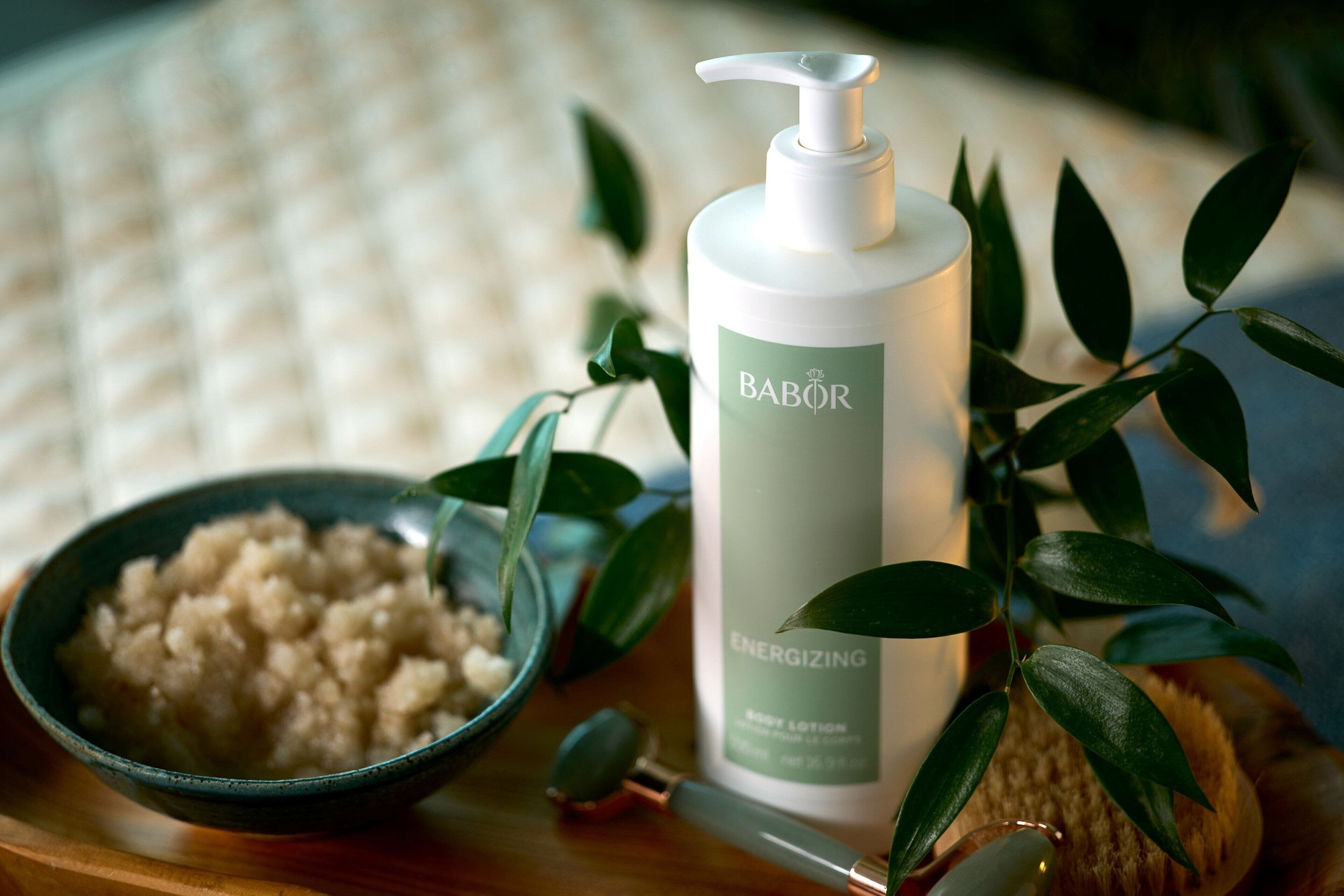 A bottle of Babor Energizing body lotion, a bowl of bath salts, and a green plant on a wooden surface.