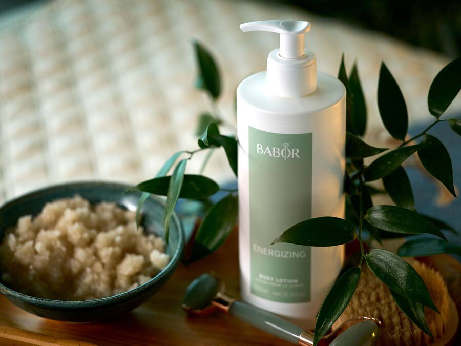 A bottle of Babor Energizing body lotion, a bowl of bath salts, and a green plant on a wooden surface.