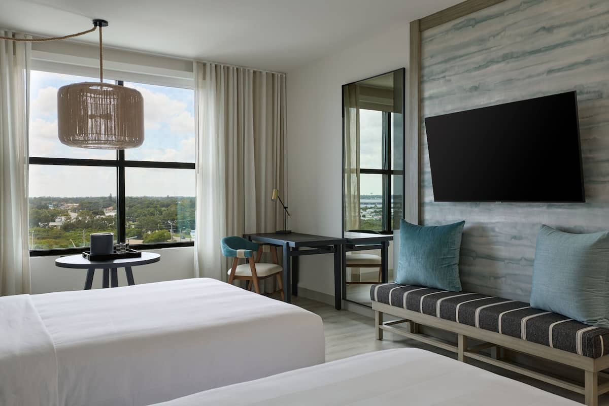 A hotel room with two beds, a desk, a chair, a sofa, a TV, and a large window with a view.