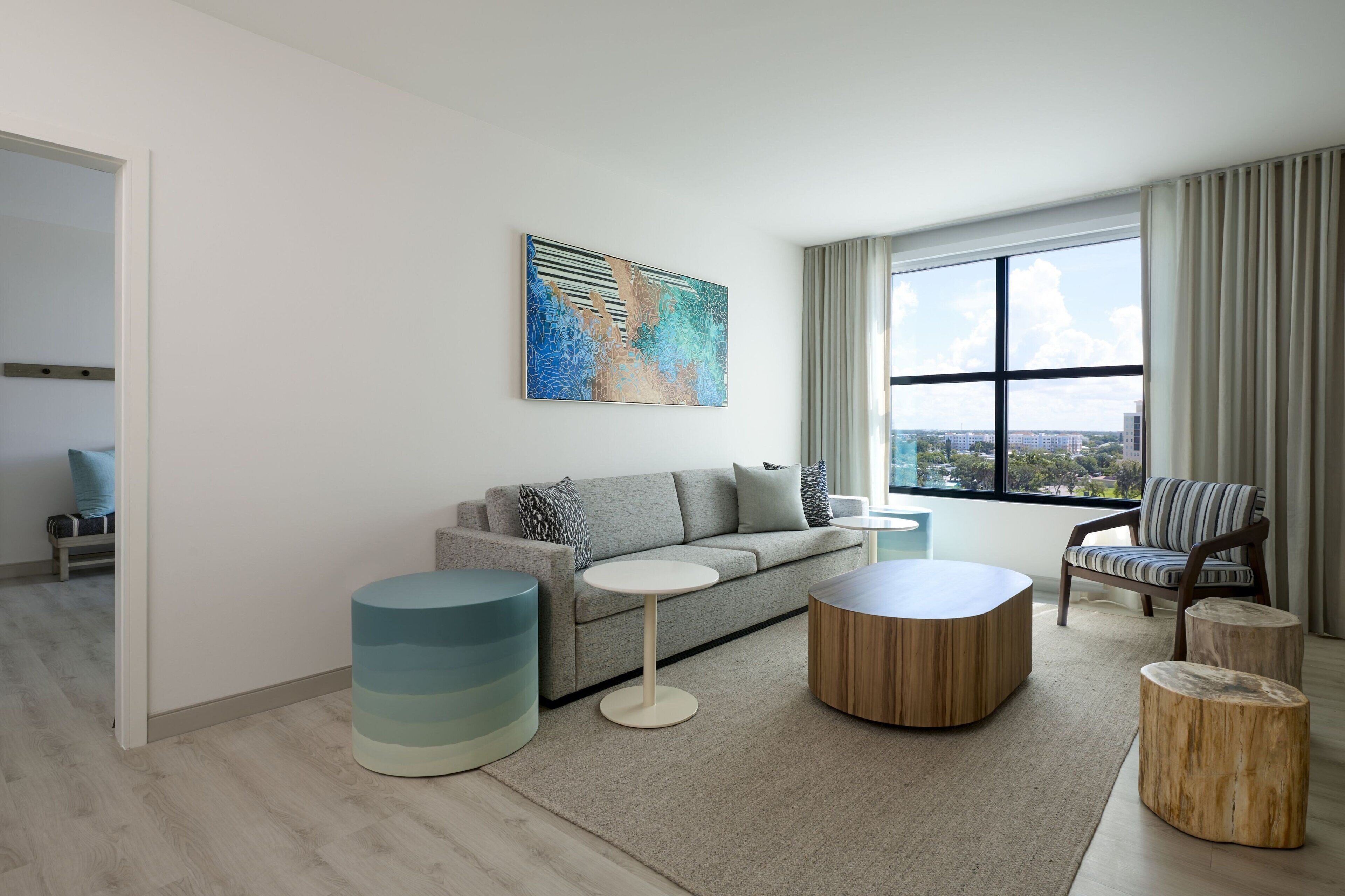 Un salón moderno con un sofá gris, una mesa de centro redonda y un sillón a rayas. Un cuadro abstracto grande adorna la pared, y hay una ventana con cortinas.