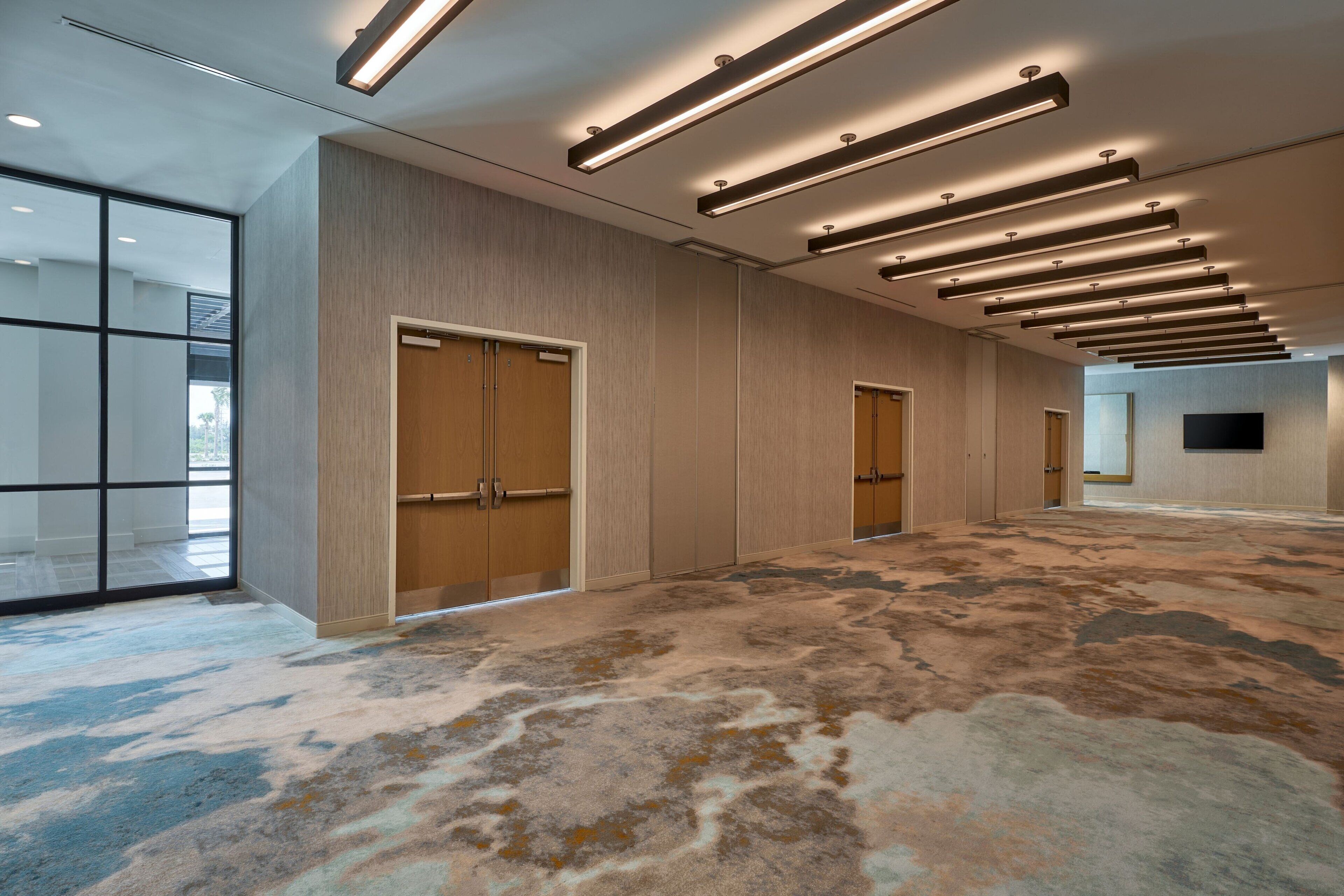 A spacious hotel lobby with a patterned carpet, wooden doors, and modern lighting fixtures.
