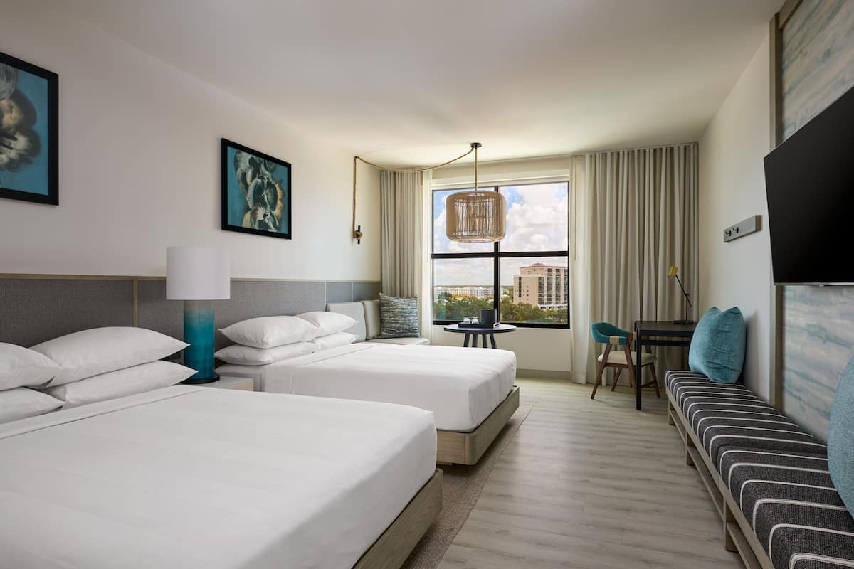 A hotel room with two beds, a sofa, a desk, and a window with a city view.