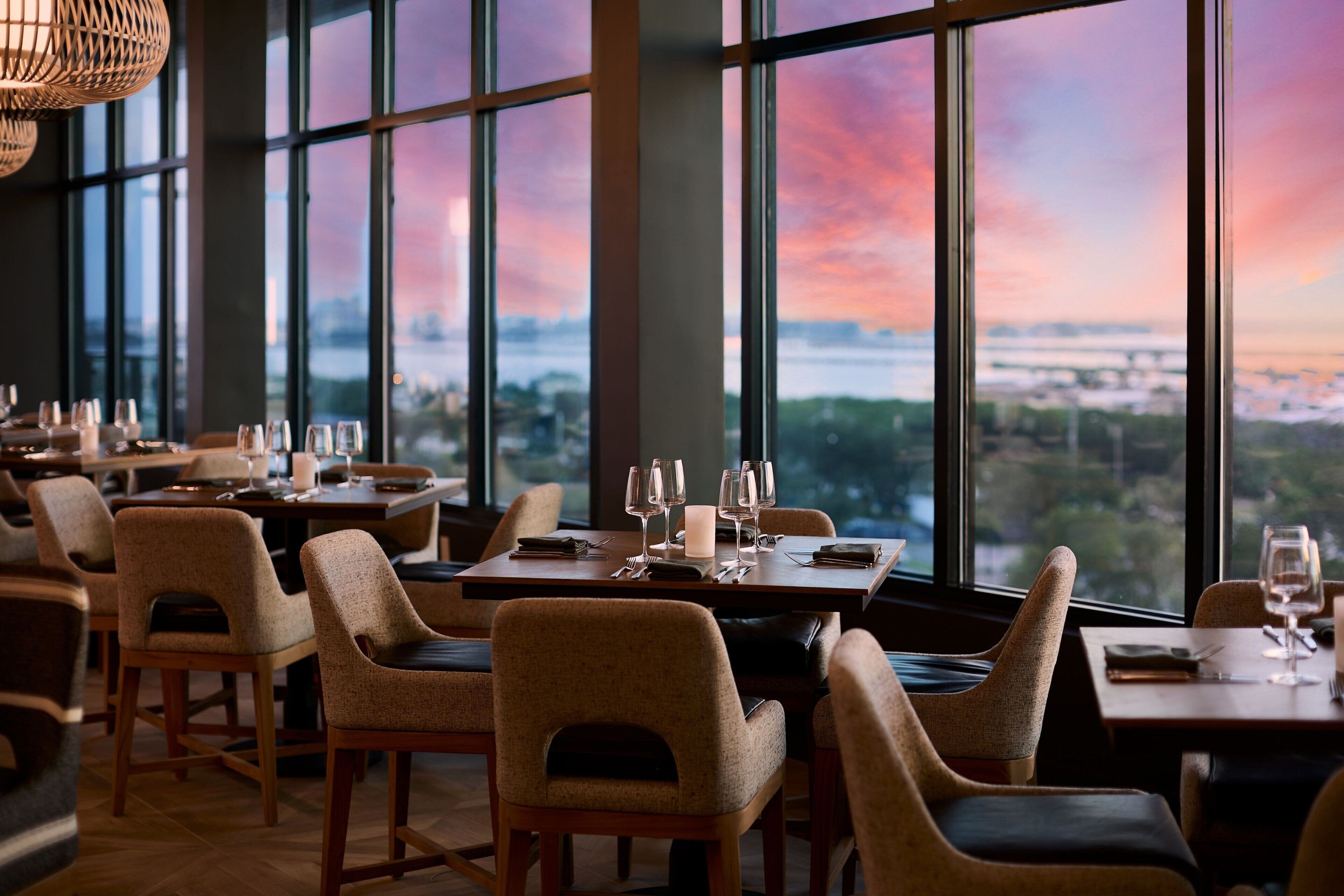 Un restaurante con mesas dispuestas para comer, ventanas amplias y vistas a un paisaje urbano.