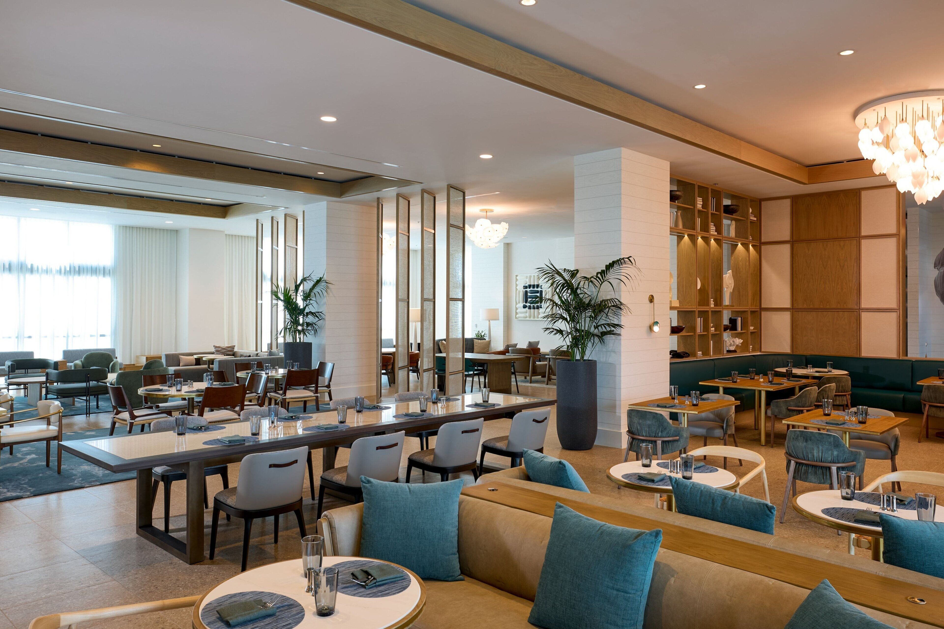 A modern hotel lobby with a mix of seating areas, including a long communal table, individual tables, and sofas. There are potted plants and a large bookshelf in the background.