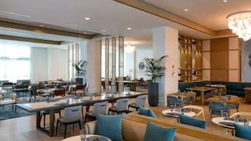 A modern hotel lobby with a mix of seating areas, including a long communal table, individual tables, and sofas. There are potted plants and a large bookshelf in the background.