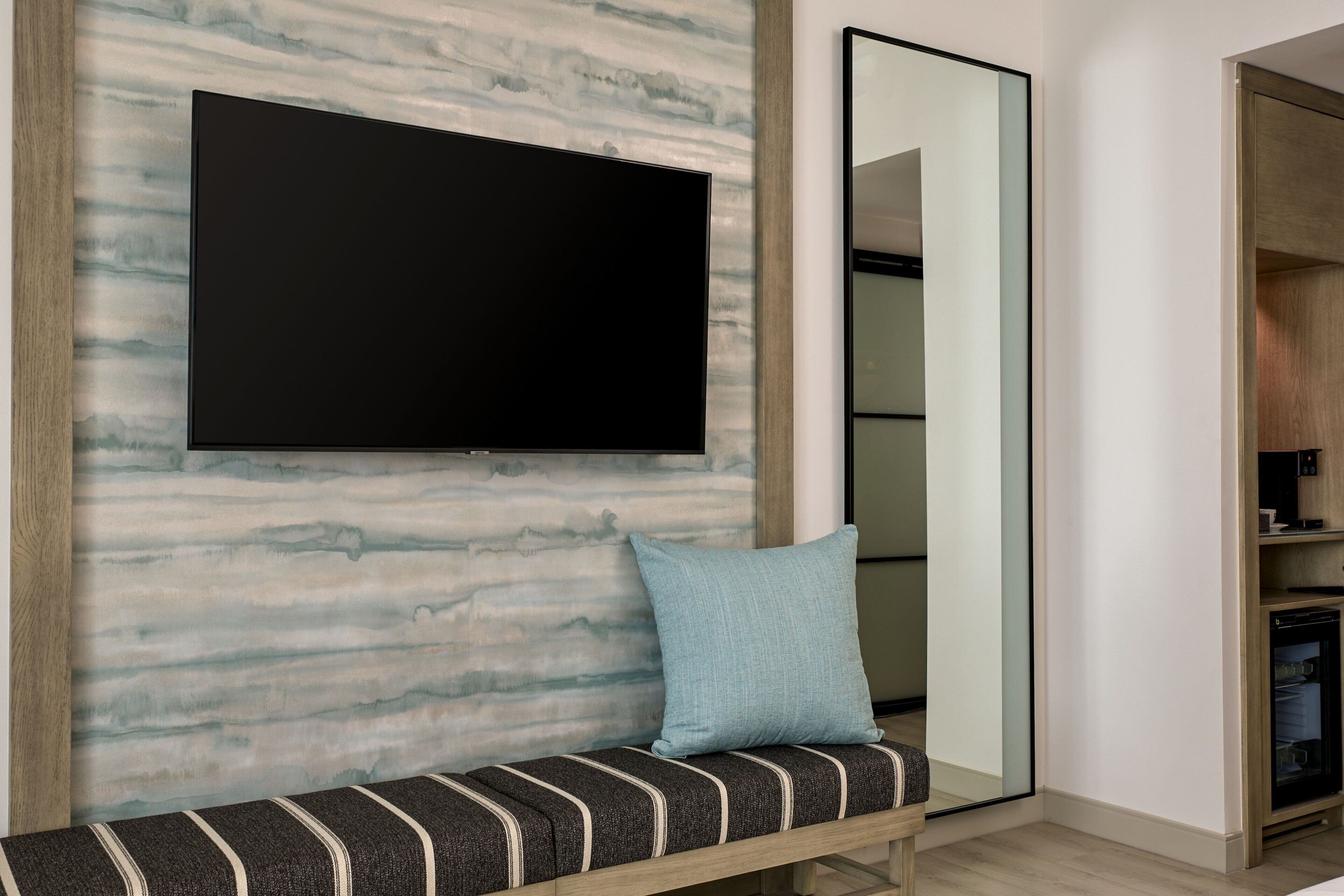 Una camera moderna con una grande TV a schermo piatto fissata a una parete con decoro marmorizzato, una panca con cuscini a righe e uno specchio che riflette l'ambiente.