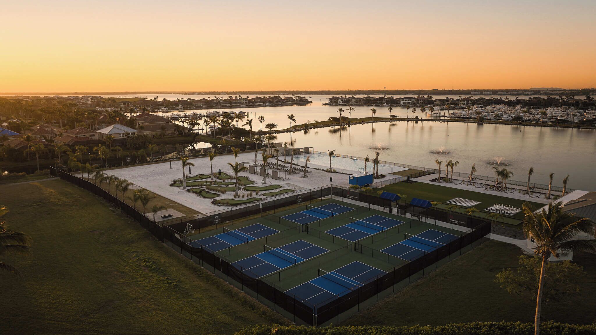 A tennis court complex with multiple courts, a pool, and a marina in the background.