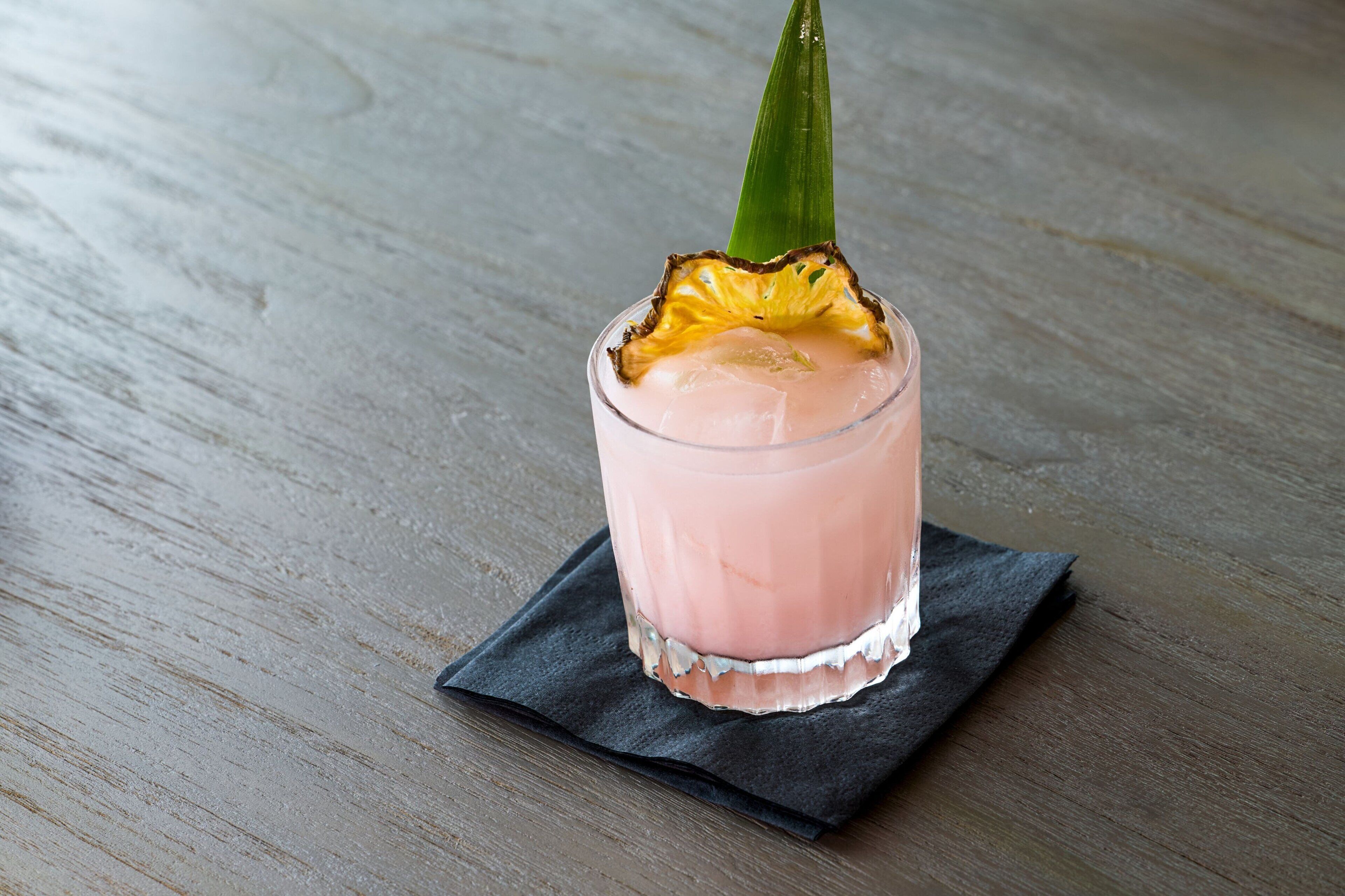 A glass of pink beverage with a pineapple garnish on a dark napkin, placed on a wooden surface.
