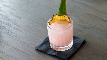 A glass of pink beverage with a pineapple garnish on a dark napkin, placed on a wooden surface.