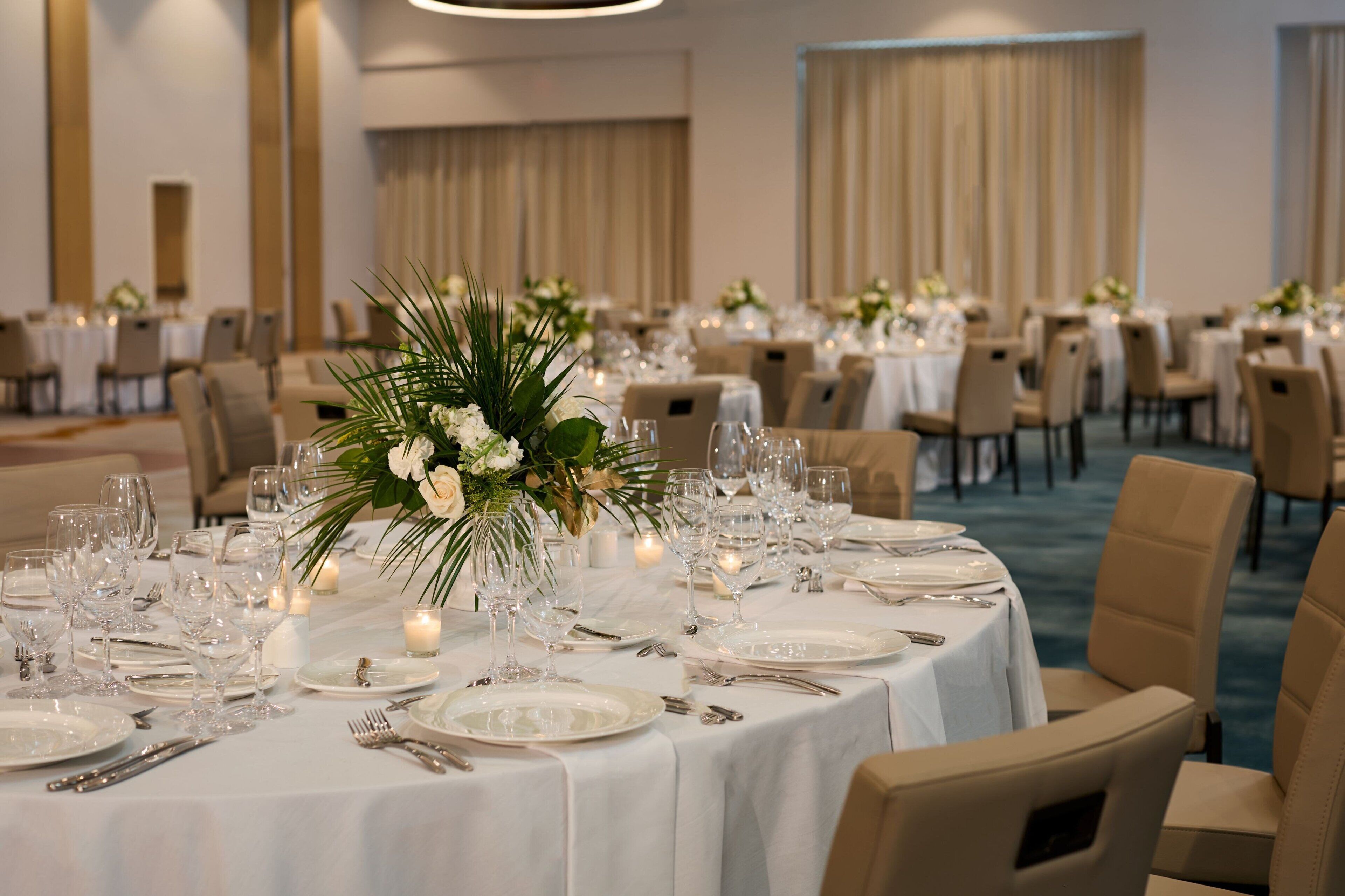 Un salón de banquetes con mesas redondas dispuestas para una comida, con manteles blancos, cristalería y centros de mesa florales.
