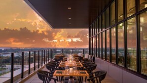 A dining area with a long table set for six, surrounded by wicker chairs, large windows, and a view of the sunset over a cityscape. - Palmetto Marriott Resort & Spa (Palmetto)