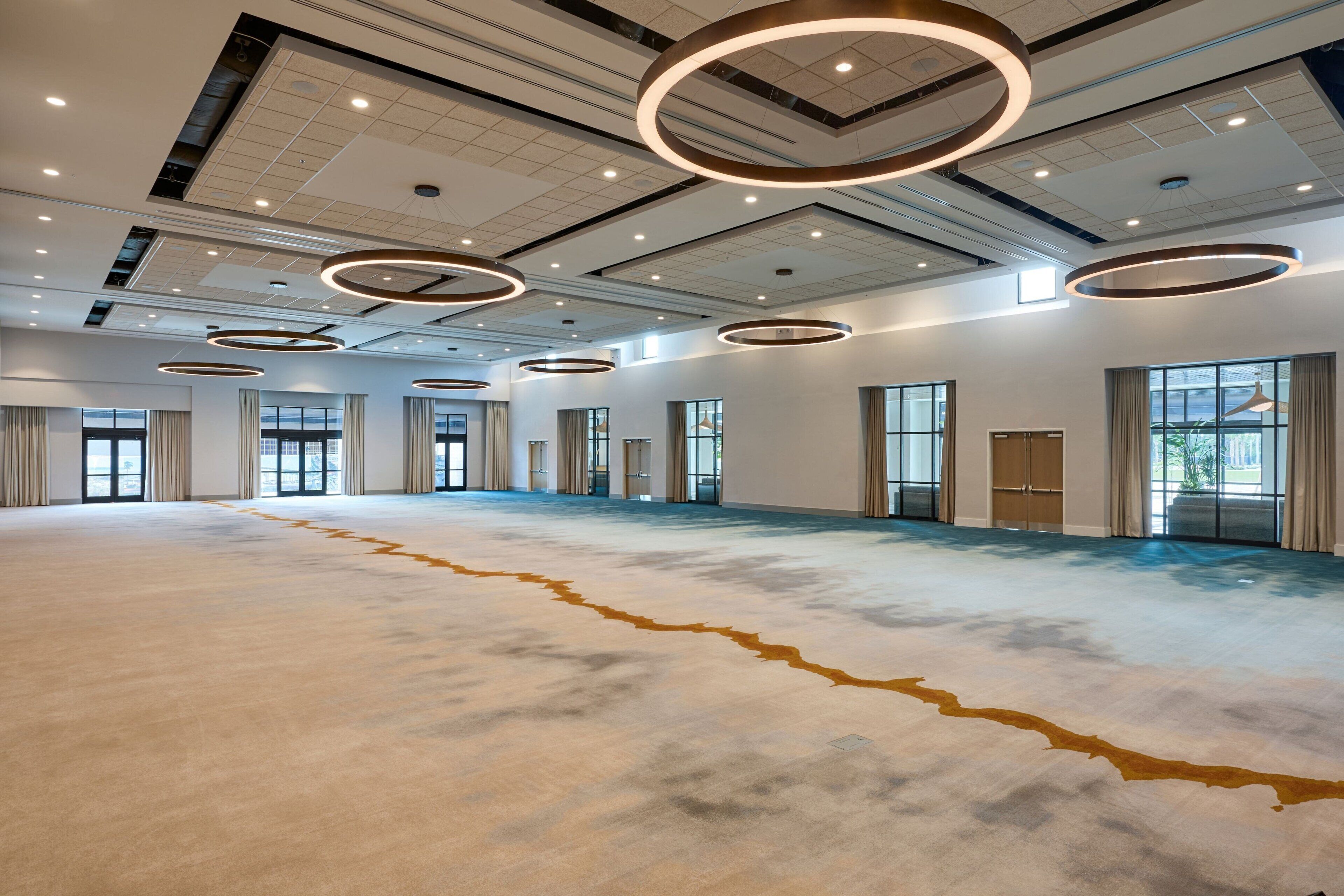 A spacious hotel lobby with a high ceiling, circular ceiling lights, large windows, and a carpeted floor.