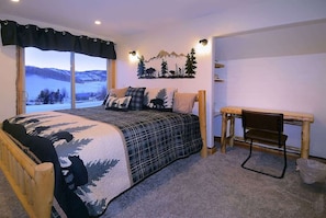A bedroom with a large window, a bed with a mountain-themed blanket, a desk, and a chair.