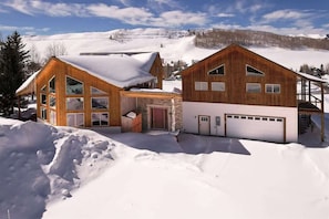 A wooden cabin with large windows, a garage, and a snow-covered landscape.