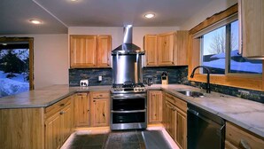 A modern kitchen with wooden cabinets, a stainless steel range hood, a dishwasher, and a window with a view of a snowy landscape.