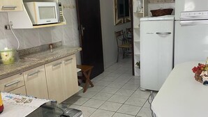 Una cocina compacta con estufa a gas, microondas y refrigerador blanco. Cuenta con una mesa de comedor con sillas y un taburete pequeño de madera. La cocina tiene gabinetes de color claro y piso de baldosas.