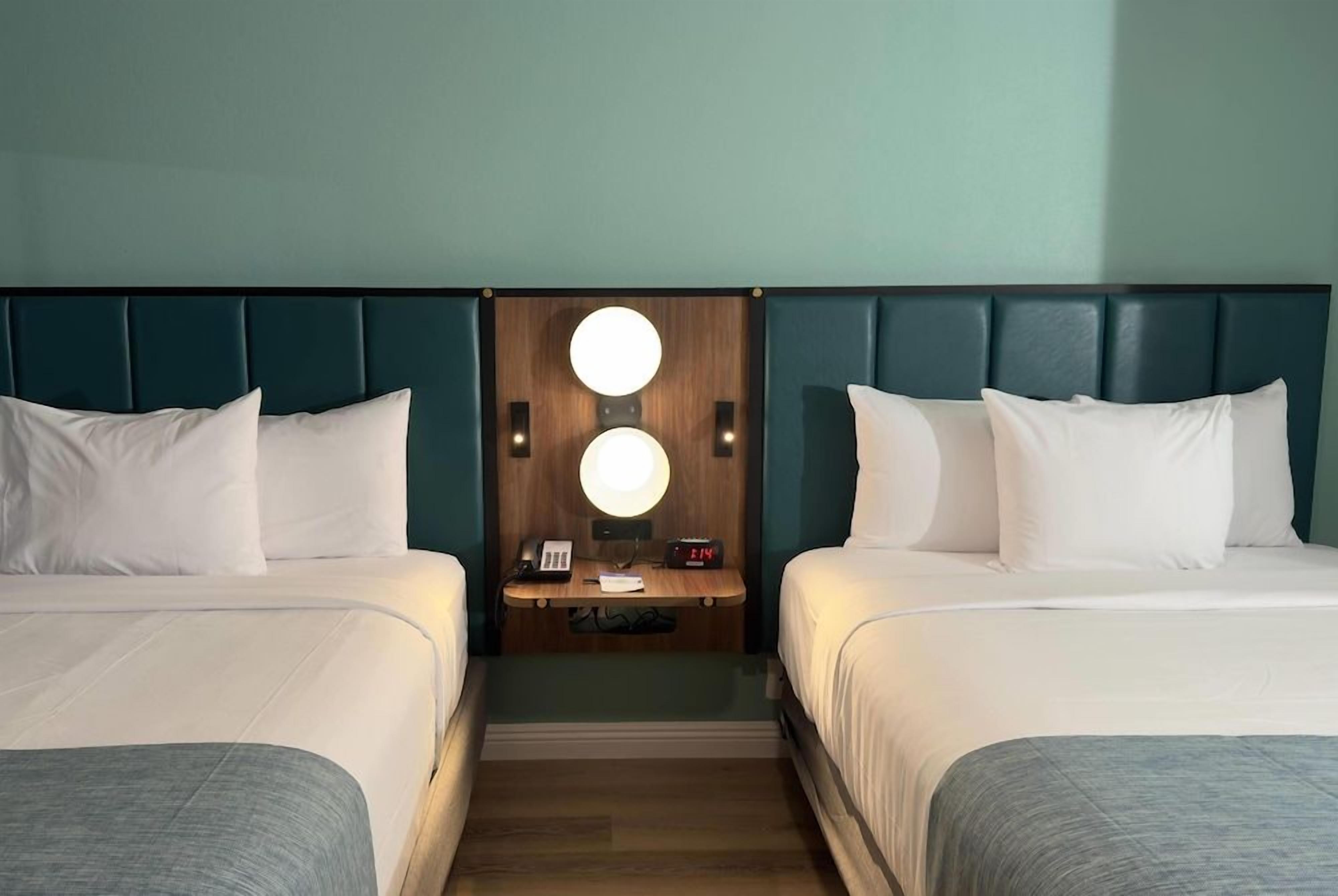 Habitación de hotel con dos camas, cabecero de madera, mesita de noche con lámpara y un reloj.