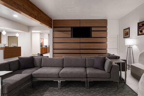 A modern living room with a sectional sofa, a flat-screen TV mounted on a wooden panel wall, a small side table, and a floor lamp. - Wingate by Wyndham Farmington NM (Farmington)