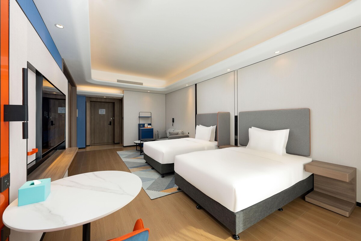 A modern hotel room with two beds, a small table, and a seating area.