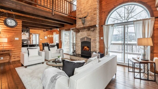 L’intérieur d’une cabane en rondins, avec une cheminée, des meubles en bois, un coin repas et une grande fenêtre.