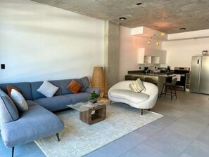Un salon moderne avec un canapé gris, un fauteuil blanc et une table basse. Il y a également un coin cuisine équipé d’un réfrigérateur, d’une cuisinière et d’un espace repas.