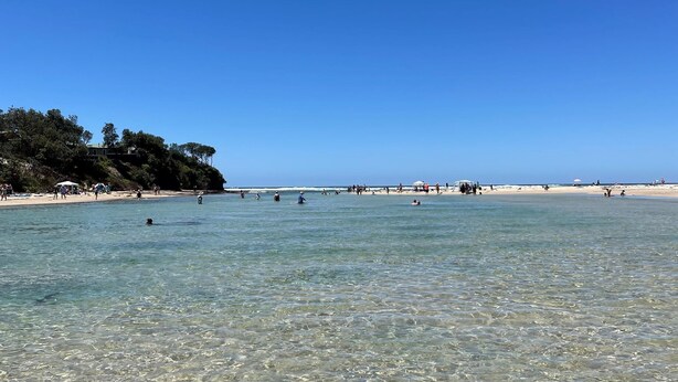 Una platja amb aigües poc profundes i cristal·lines, gent nedant i prenent el sol, i un penya-segada amb vegetaci ó al fons.