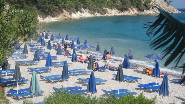 Une plage bordée de rangées de transats et de parasols bleus, un littoral rocheux et une mer d’un bleu limpide.