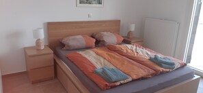 Una cama bien tendida con un edredón a rayas y dos almohadas, una mesita de noche con una lámpara, y una ventana con persianas.